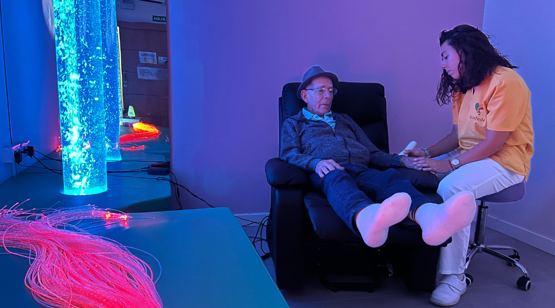 SPAP Centro Un hombre mayor está sentado en una silla reclinable en una sala de estimulación sensorial, acompañado por una trabajadora de una residencia.